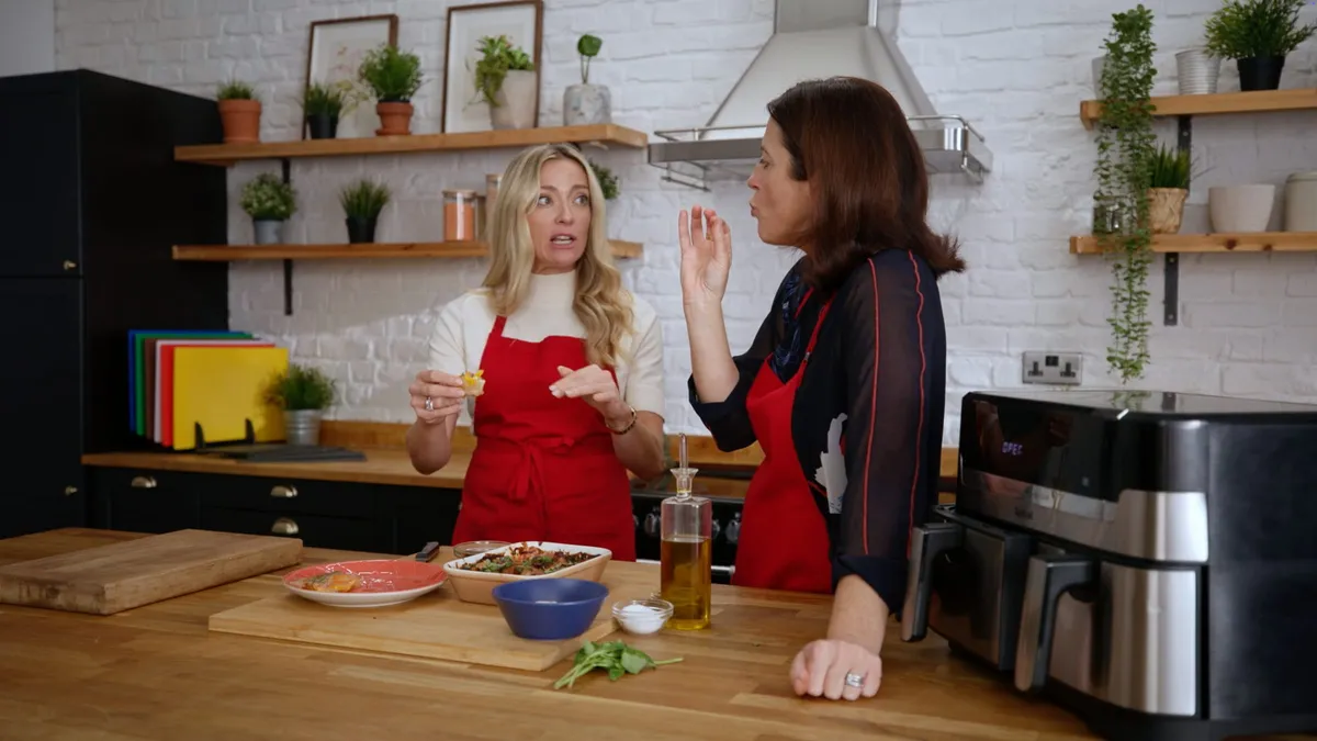 A blonde woman in a red apron and a dark-haired woman in a black top and red apron talk over a wooden table with food and an air fryer, in a white-brick kitchen.