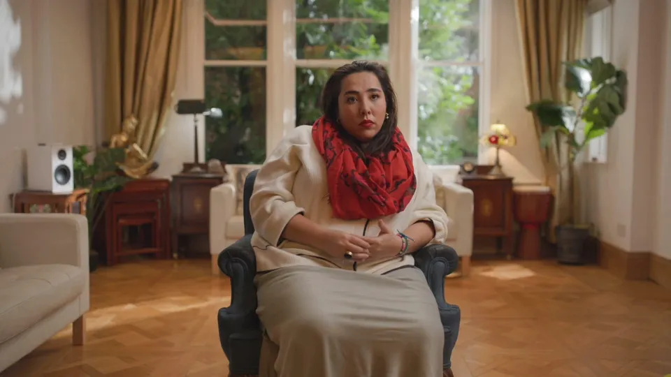 A contributor wearing a red scarf sits in an armchair in a sitting room, facing camera, daylight coming through windows behind her.