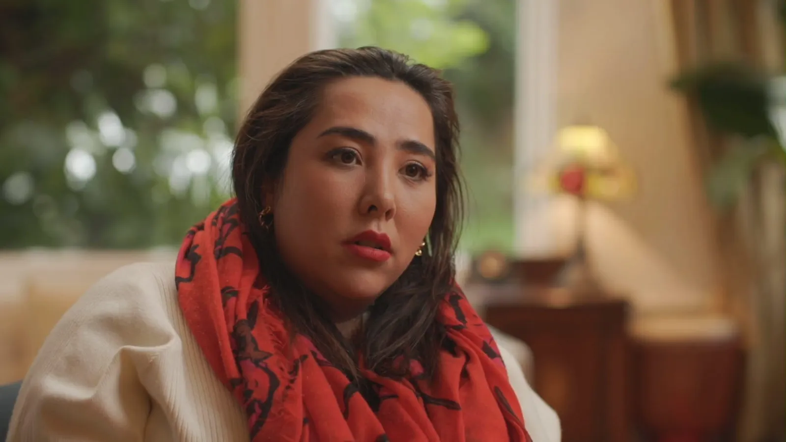 Close-up of a contributor wearing a red patterned scarf and cream jacket, seated indoors, a window and lamp softly blurred behind her.