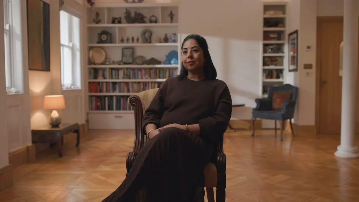 A contributor sits in a wooden armchair in a large room, with bookshelves and a lit table lamp visible behind her.