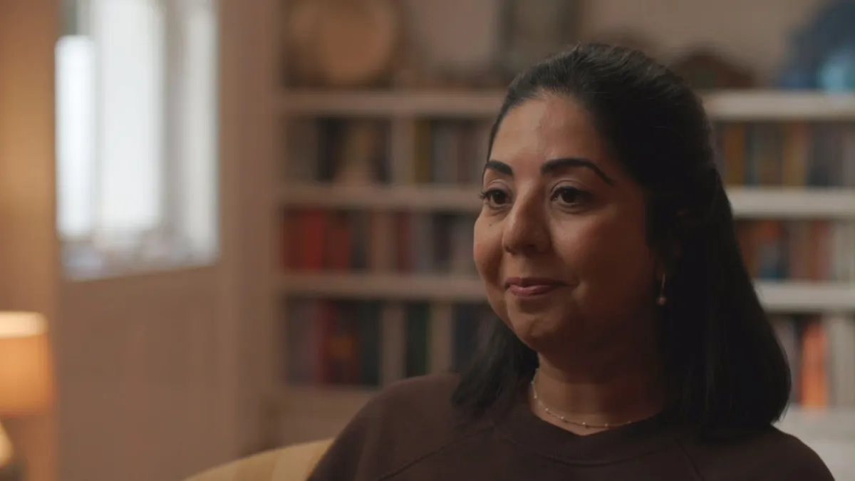 Close-up of a contributor seated indoors, looking off-camera, with bookshelves and a window behind her lit by soft daylight.