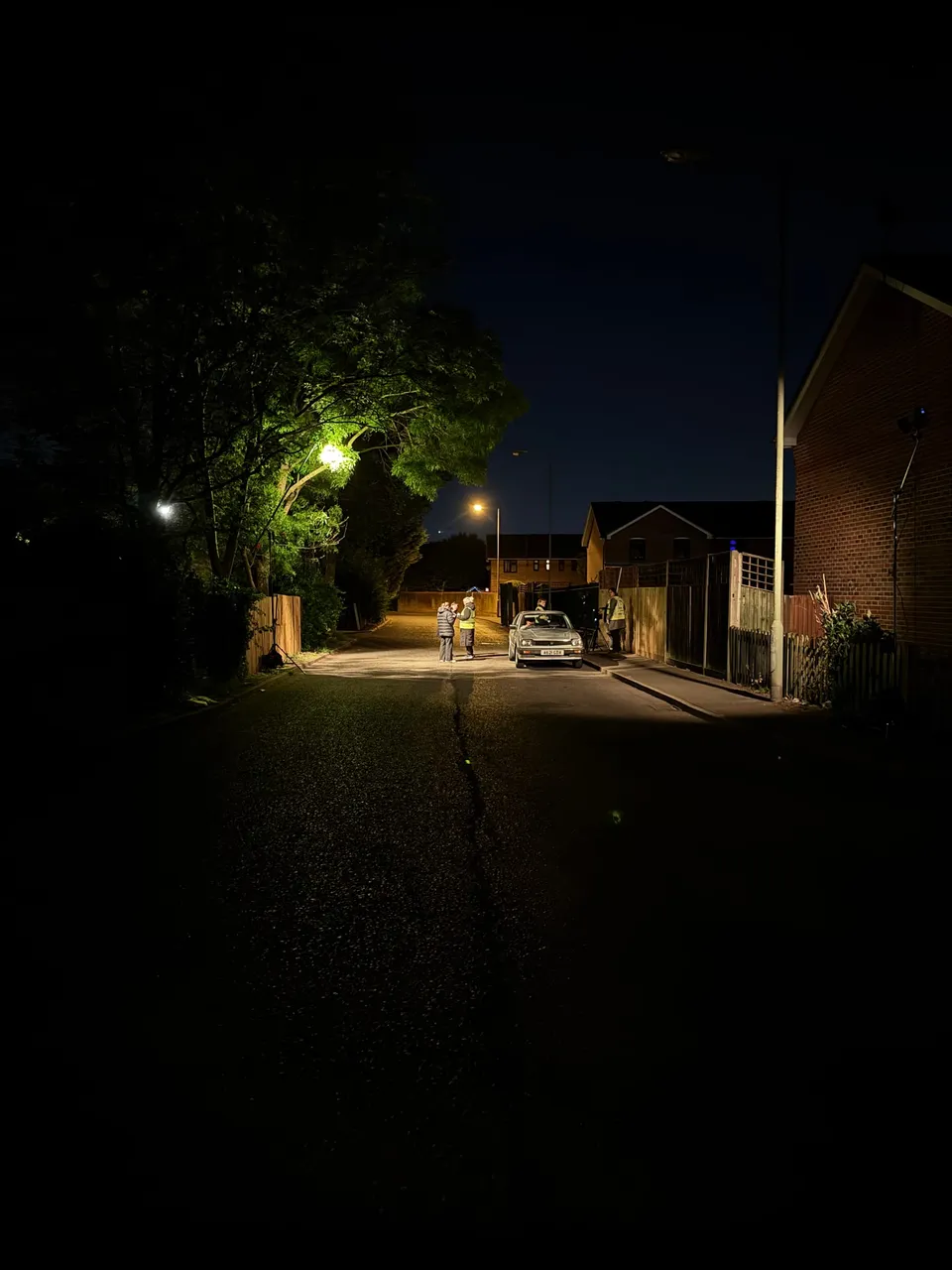 Night-time residential street lit by a streetlamp, with a small crew and actors gathered around a parked saloon car, houses and trees in shadow beyond.