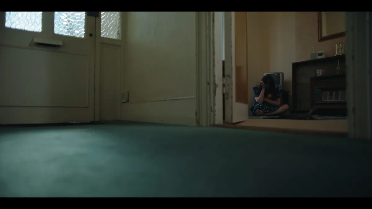 Hallway with a glass-panelled front door, seen beyond an open doorway a girl in a floral dress sits on the floor against a fireplace, head in her hand.