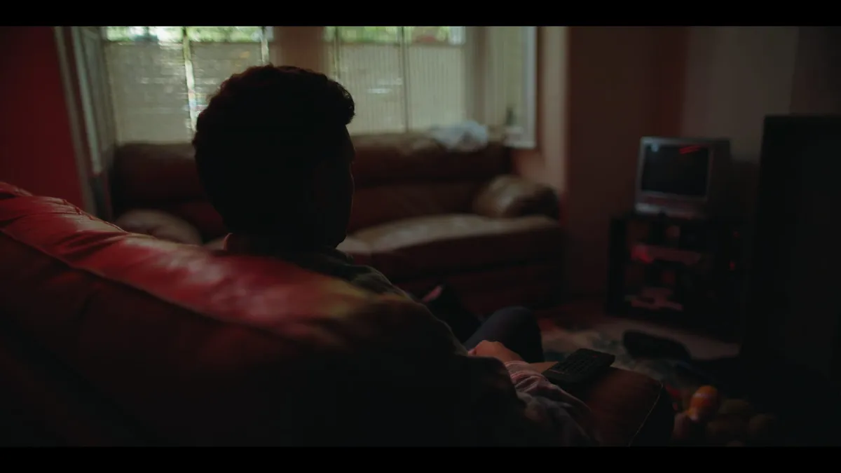 A man seen from behind, seated on a leather sofa in a dim living room, facing a window with net curtains and a small television.