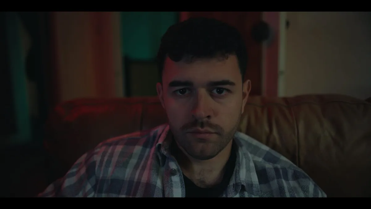 Close-up of a young man in a checked shirt sitting on a leather sofa, facing camera in dim, red-tinted light.