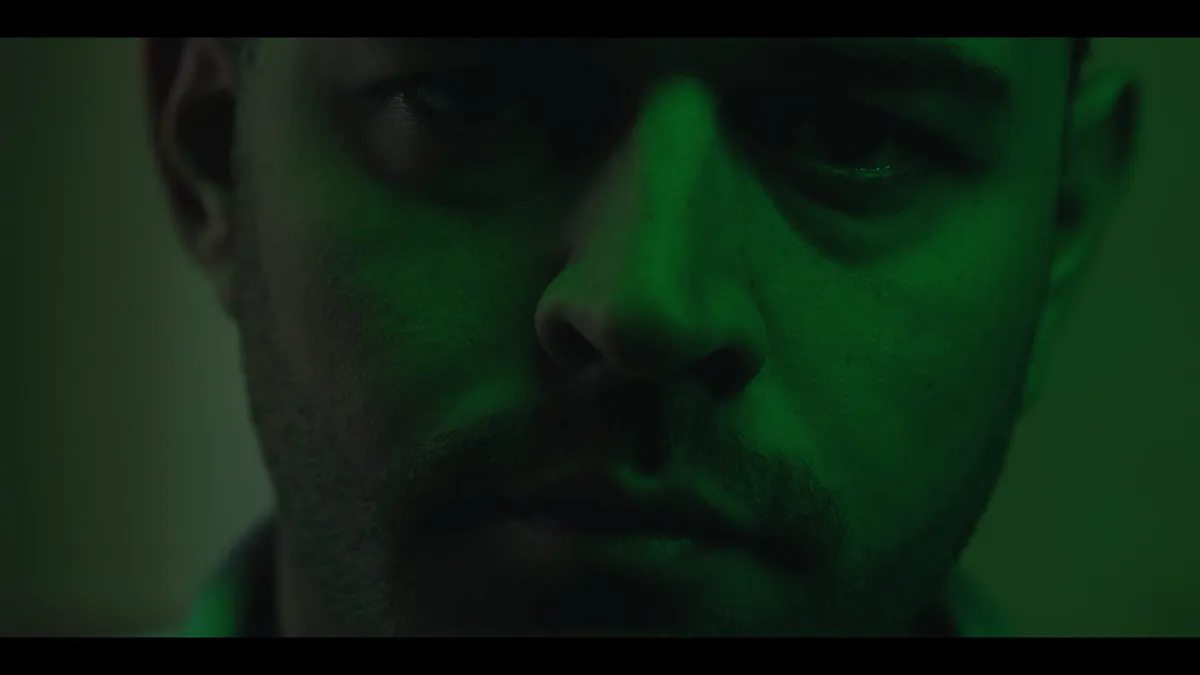 Extreme close-up of a man's face bathed in green light, eyes downcast, looking off-camera.