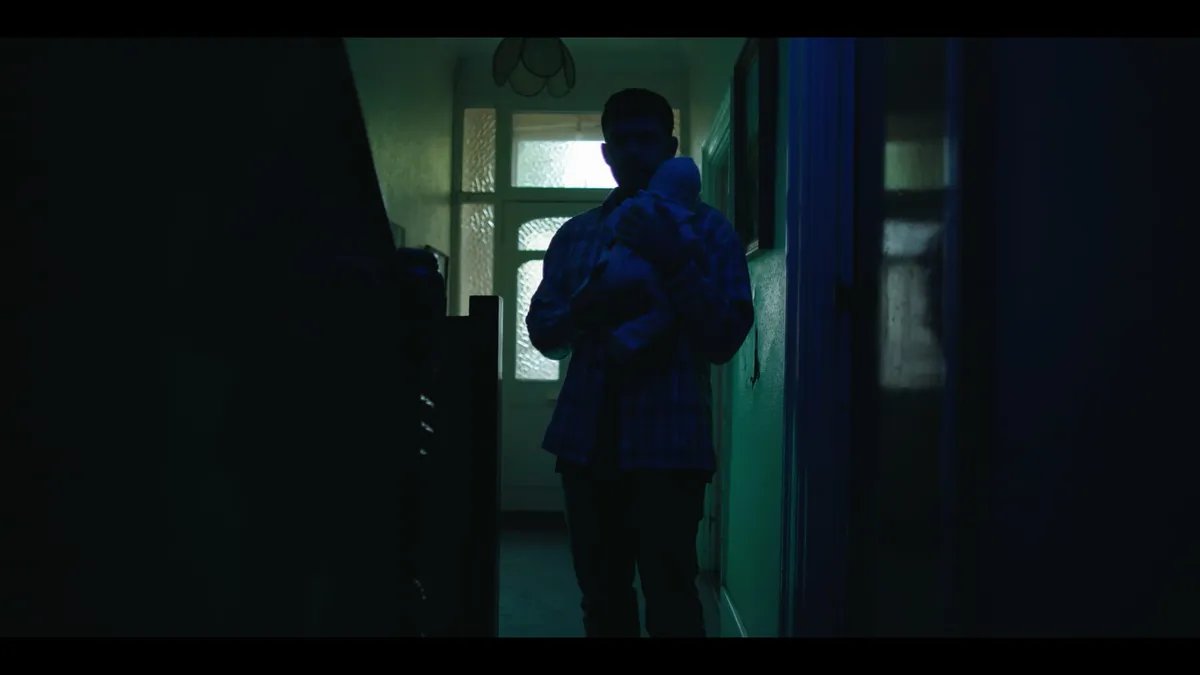 A man in a checked shirt stands in a dim hallway holding a swaddled infant, facing the camera, lit by a glazed front door behind him.