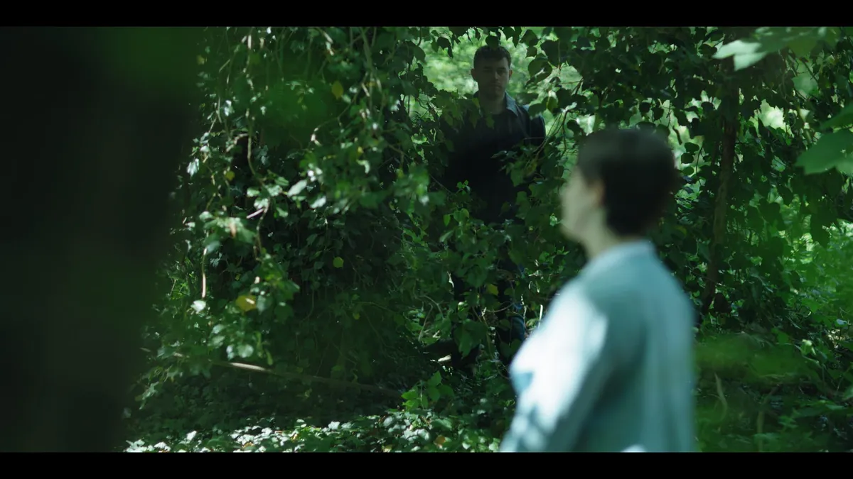 A man stands among dense woodland foliage, facing camera, while a woman with short hair looks towards past him in the foreground, out of focus.