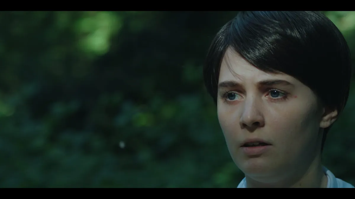 Close-up of a woman with short dark hair, positioned right of frame, looking off-camera with a startled expression against a blurred green woodland background.