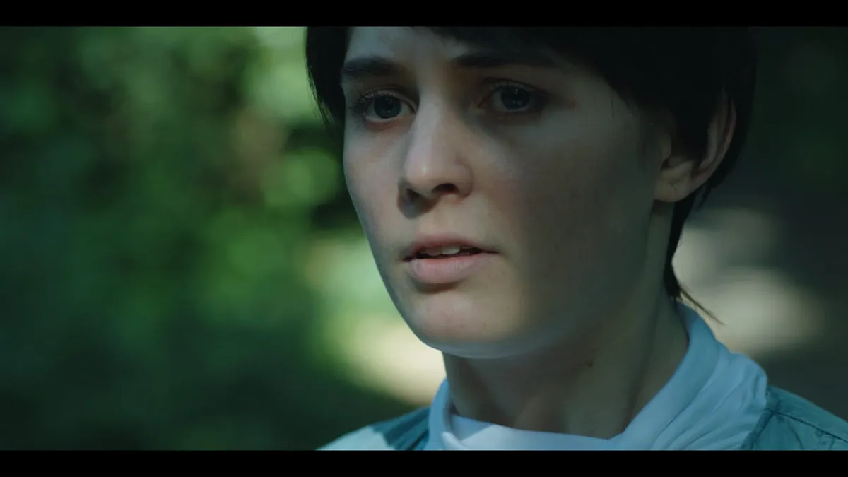 Close-up of a young woman with short dark hair, mouth slightly open, looking off-camera, against a blurred green woodland background.