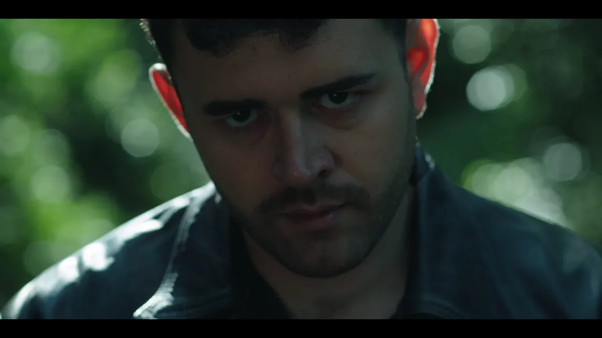 Close-up of a man in a dark leather jacket, looking down and away from camera, backlit foliage blurred behind him.