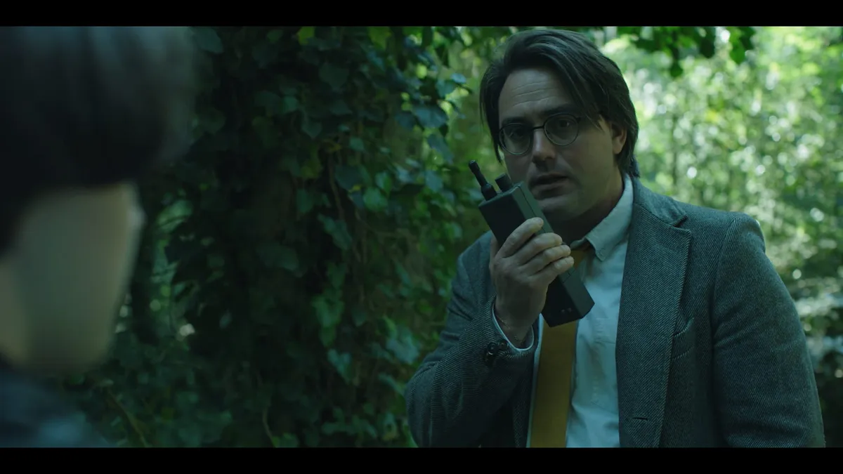 A man in a tweed jacket and yellow tie speaks into a walkie-talkie, facing an out-of-focus figure in the foreground, woodland behind him.