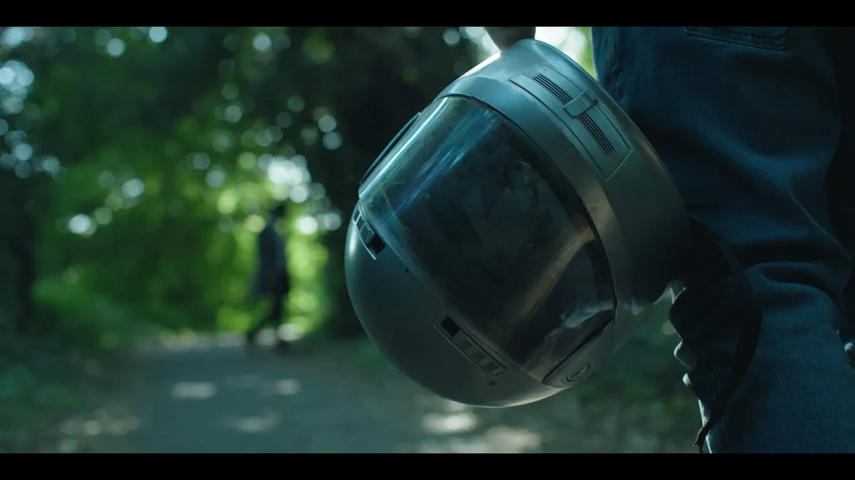A motorcycle helmet held at a person's side in close-up, with a figure walking away along a tree-lined path in the blurred background.
