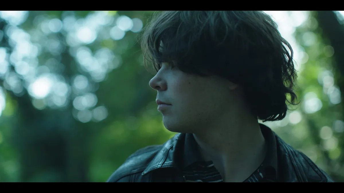 Close-up profile of a young man with tousled dark hair in a leather jacket, looking off-camera, set against a blurred, sunlit woodland background.