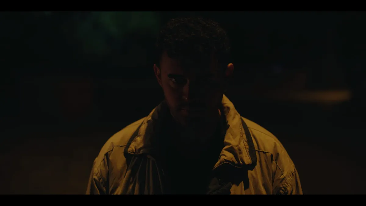 Close-up of a man in a jacket standing in near darkness, warm low light on his face as he looks down, head slightly bowed.