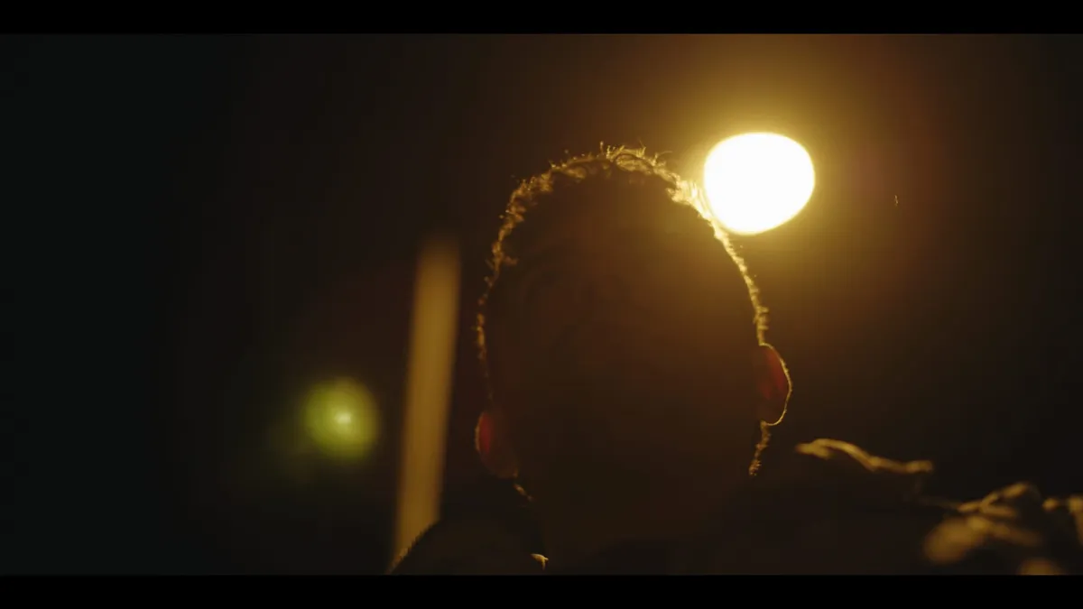 Close-up of a man's face lit from behind by a streetlight at night, features in shadow, head tilted upward.