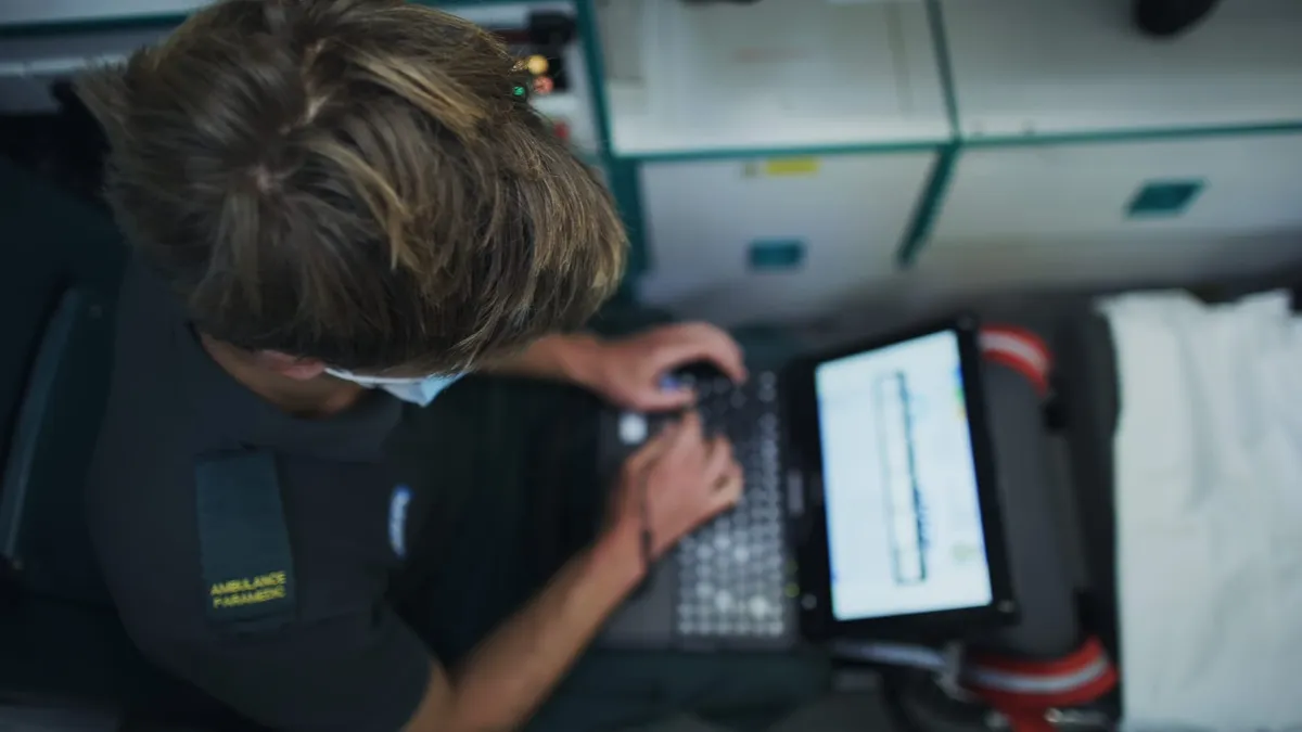 Overhead view of a paramedic with light brown hair and a face mask typing on a laptop showing a chart, "Ambulance Paramedic" patch visible.