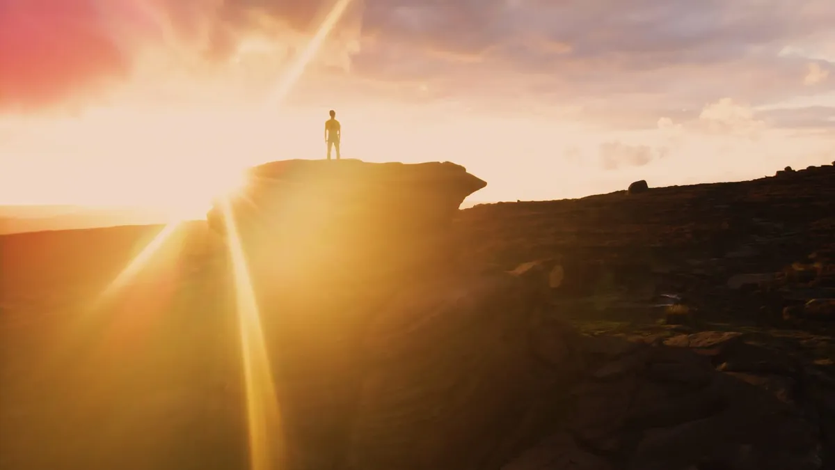 Silhouetted figure standing on a rocky outcrop, wide shot against a glowing orange and grey sunset sky, moorland stretching below.