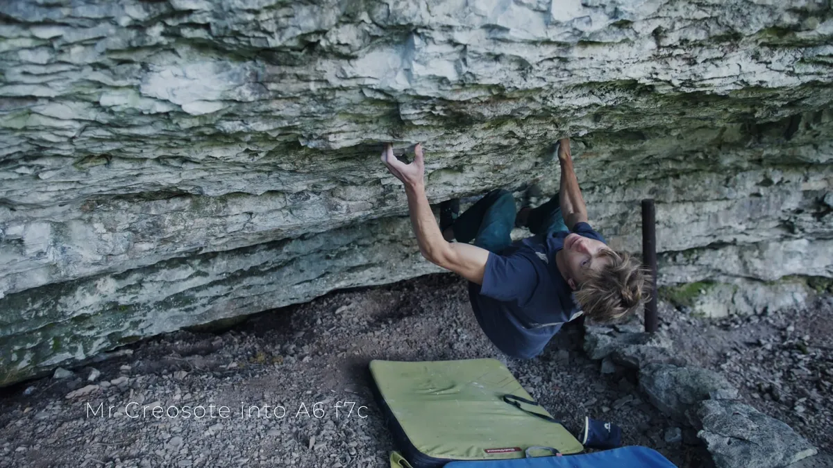 A man with light brown hair, in a dark blue t-shirt, climbs an overhanging rock face upside down, crash pad below.