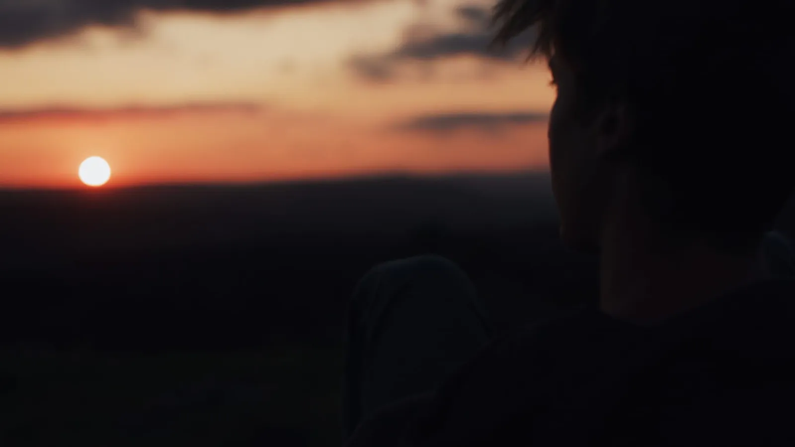 Silhouetted head and shoulders of a person in profile against a glowing orange sunset, sun low on the horizon, dark moorland below.