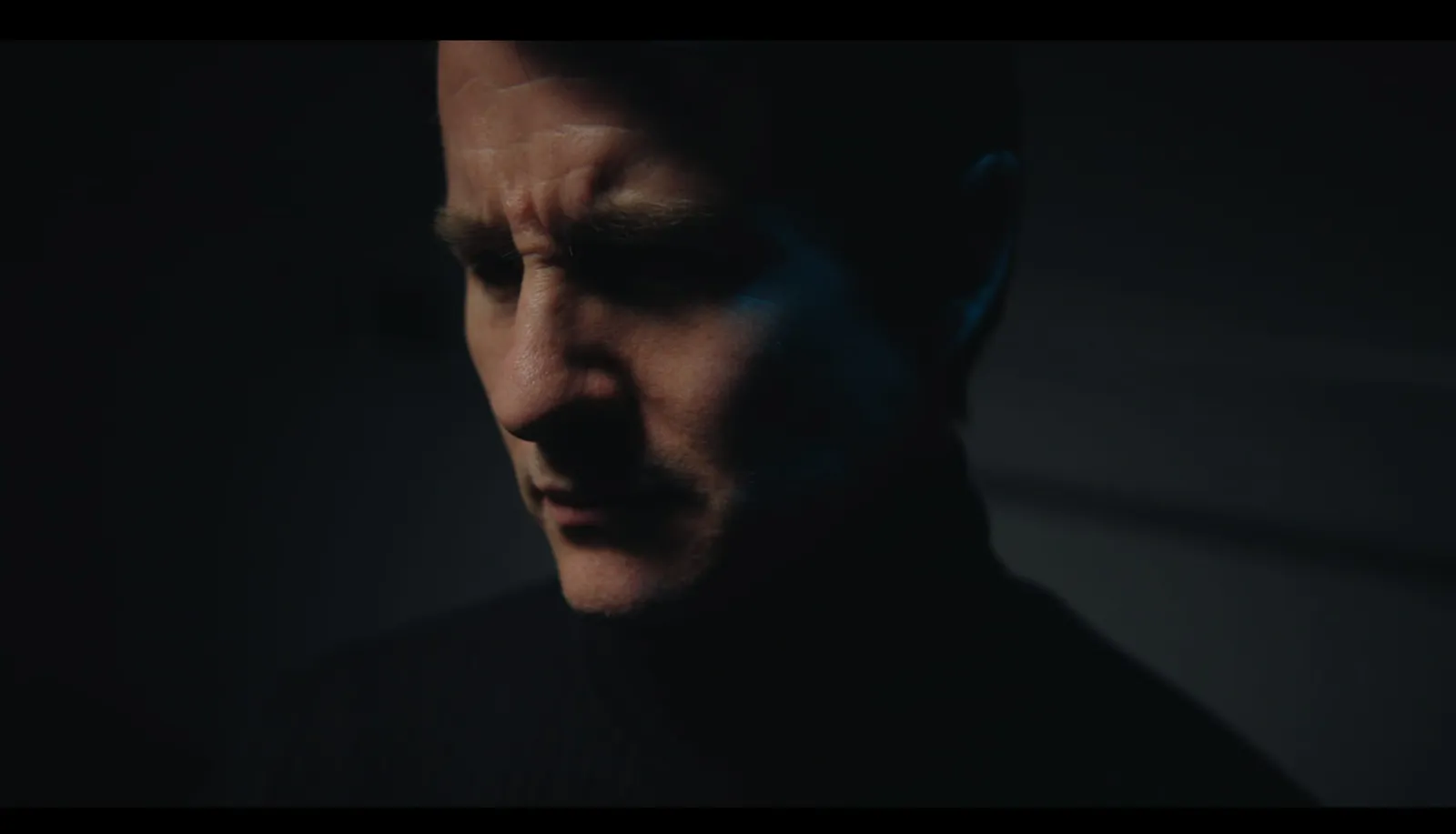 Close-up of a man in a dark polo neck, face partly lit, looking downward and away from camera in shadow.