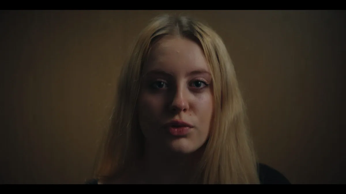Close-up of a young woman with long blonde hair, facing camera in dim light against a plain brown wall.