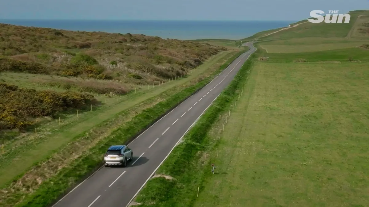 Aerial view of a silver car driving along a coastal road through green fields, with the sea visible in the distance.