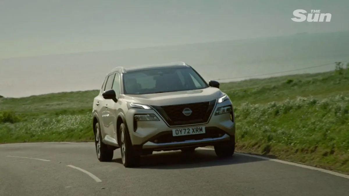 A silver SUV drives along a coastal road, sea in the background and grassy verges on either side.