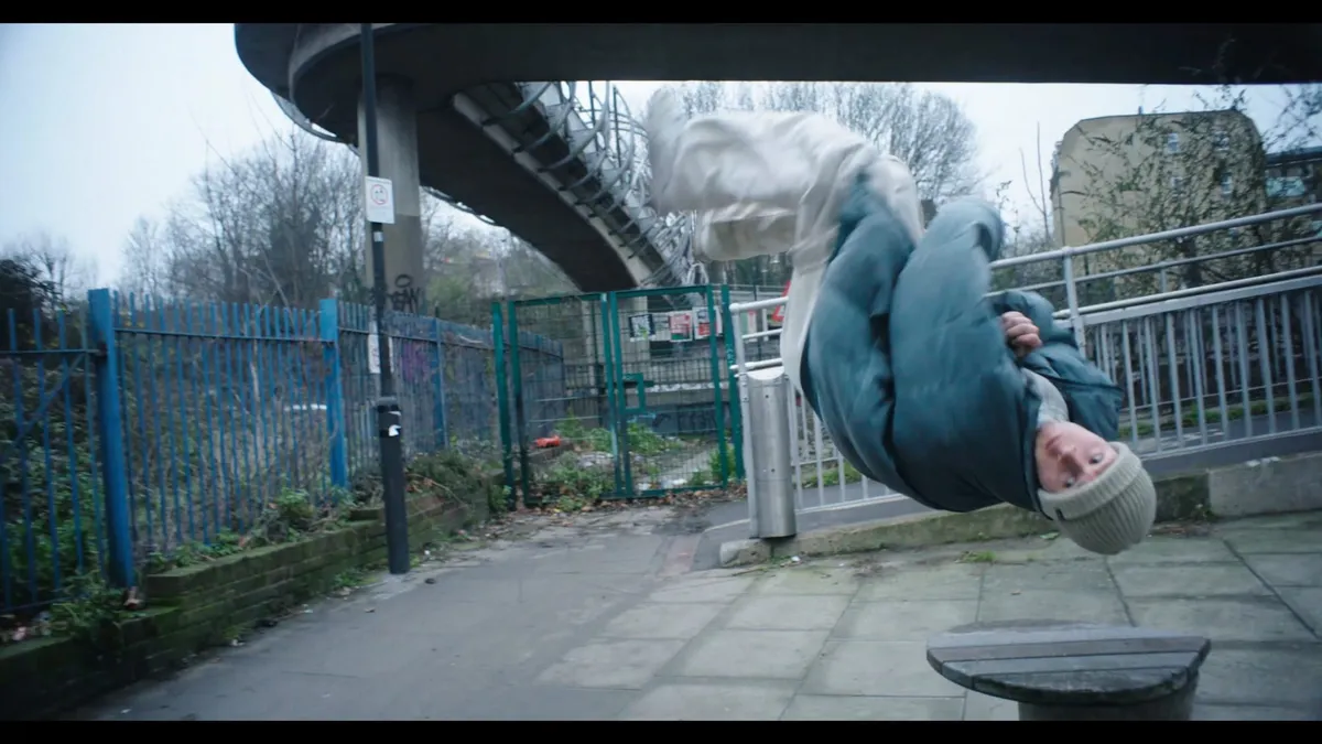 A person in a cream beanie and blue puffer jacket somersaults upside down beneath a footbridge, railings and a fenced pathway behind.