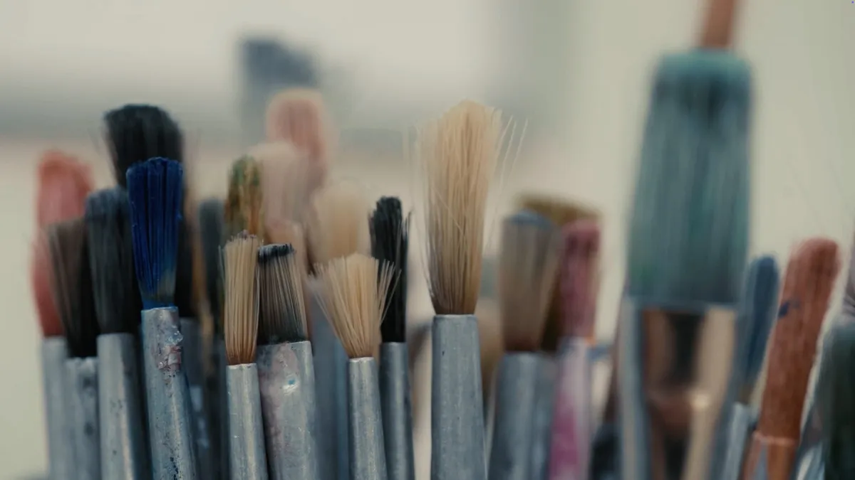 Close-up of paintbrushes with worn bristles standing upright in a holder, paint-stained handles, blurred background.