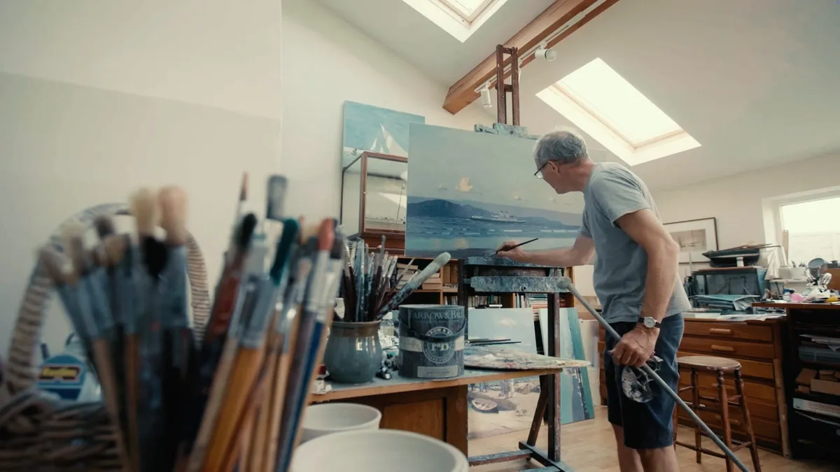 Grey-haired man in glasses and a grey T-shirt, in profile, painting at an easel in a skylit studio, paintbrushes blurred in the foreground.