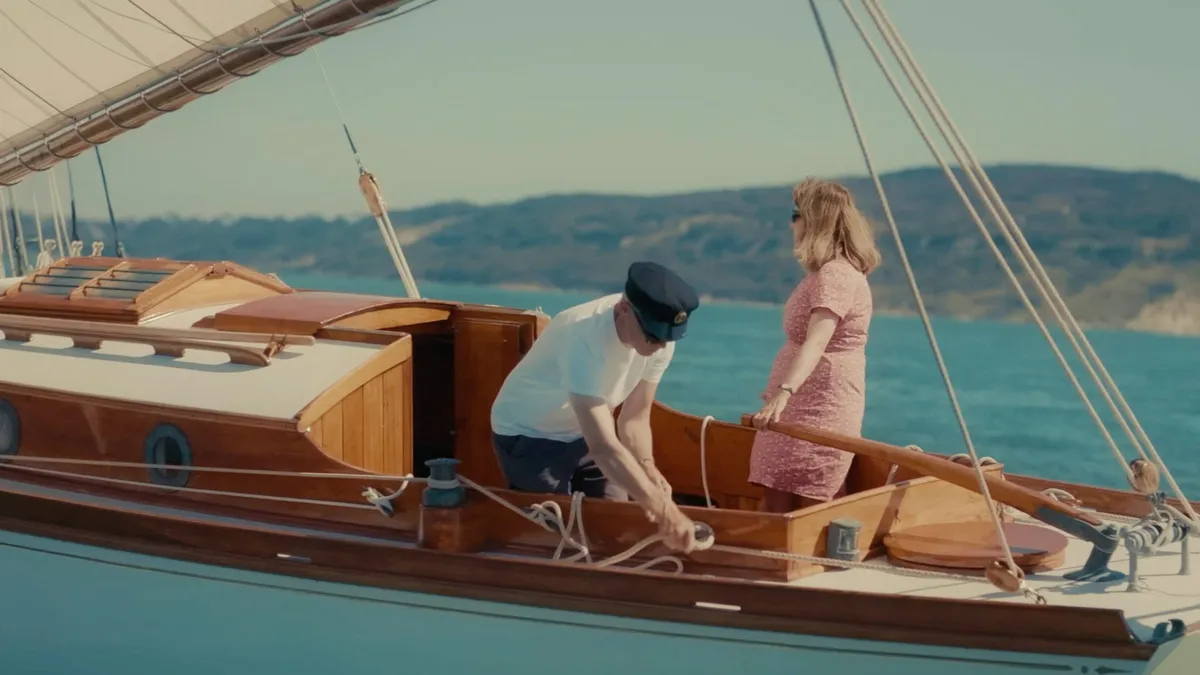 A man in a peaked cap and white T-shirt coils rope on a wooden sailing boat's deck as a woman in a pink dress stands behind him, hills across the water.