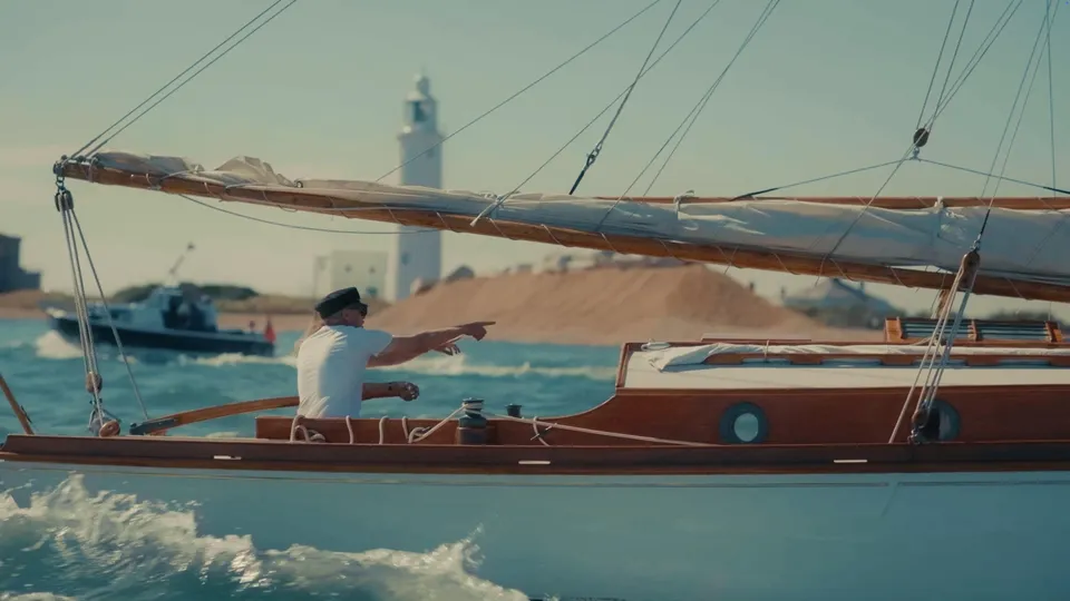 A man in a peaked cap and white T-shirt points from a wooden sailing boat, a lighthouse and small boat visible behind him on the water.