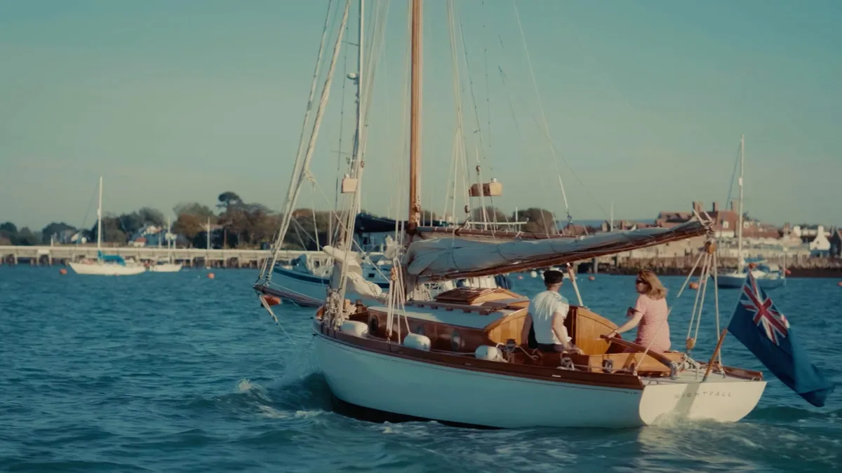 A wooden yacht with furled sail motors into harbour, a man and a woman in a pink dress at the helm, a Union flag at the stern, town buildings beyond.