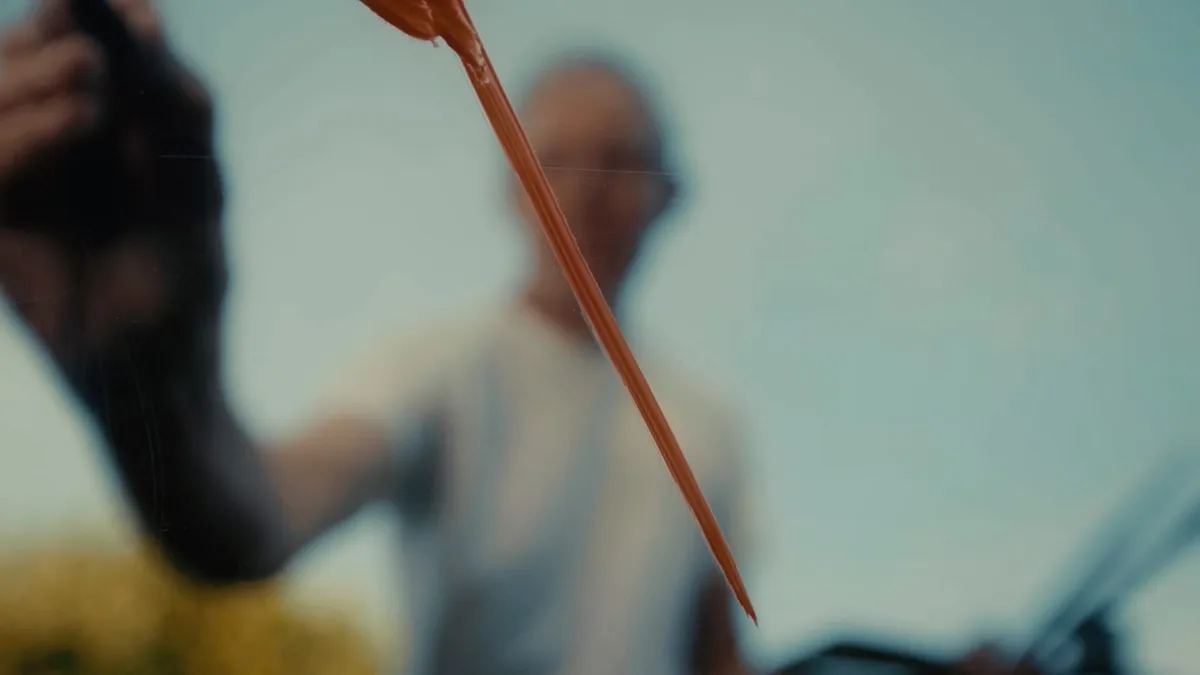 Close-up of a paint-laden brush held up against a blurred figure in glasses and a grey T-shirt in the background.