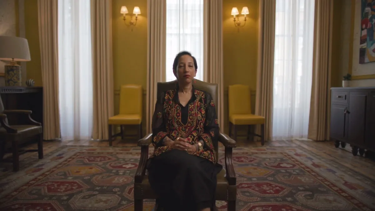 A woman with dark hair pulled back, wearing an embroidered black top, sits in a leather chair facing camera in a grand room with yellow walls.