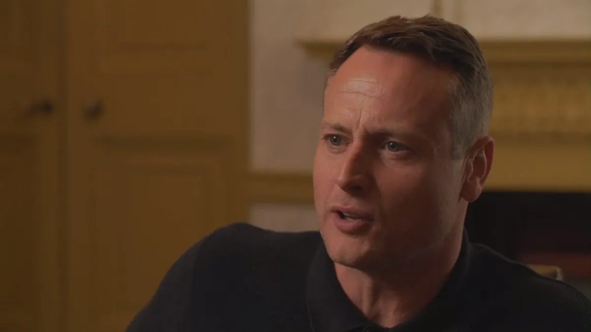 Close-up of a short-haired man in a dark polo shirt, speaking and looking off-camera, with yellow panelling and a fireplace behind him.