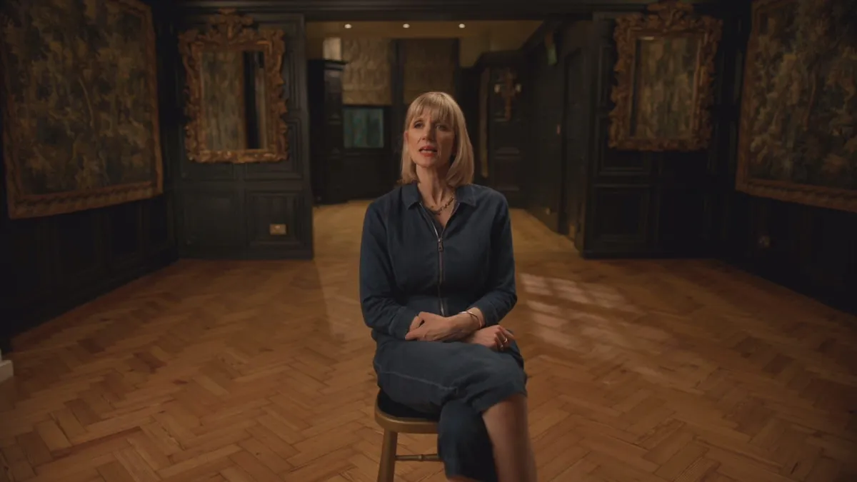 A blonde woman with a fringe, in a dark denim shirt dress, sits on a stool facing camera in a wood-panelled room with tapestries.