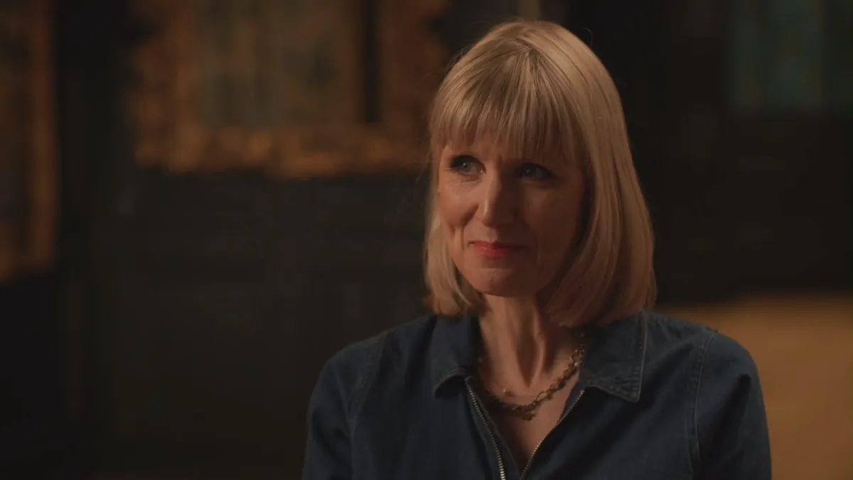 Close-up of a blonde woman with a fringe, wearing a denim shirt, looking off-camera in a dimly lit wood-panelled room.