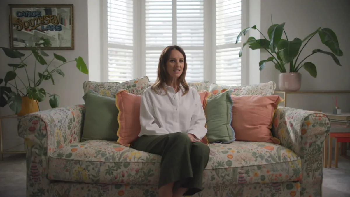 A woman with long reddish-brown hair, in a white shirt and green trousers, sits on a floral sofa facing camera in a room with shuttered windows and potted plants.