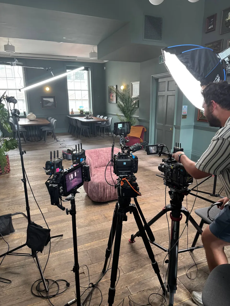 A pink armchair set up for an interview, surrounded by camera tripods and monitors, with a softbox light behind a man adjusting a camera, in a green-panelled room.