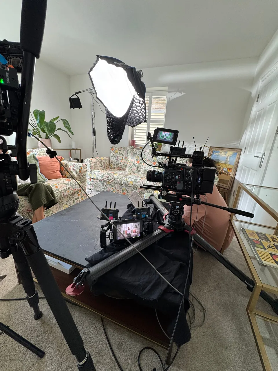 A rigged cinema camera with monitors on a slider faces a floral sofa, softbox light behind, in a domestic room with shuttered windows.