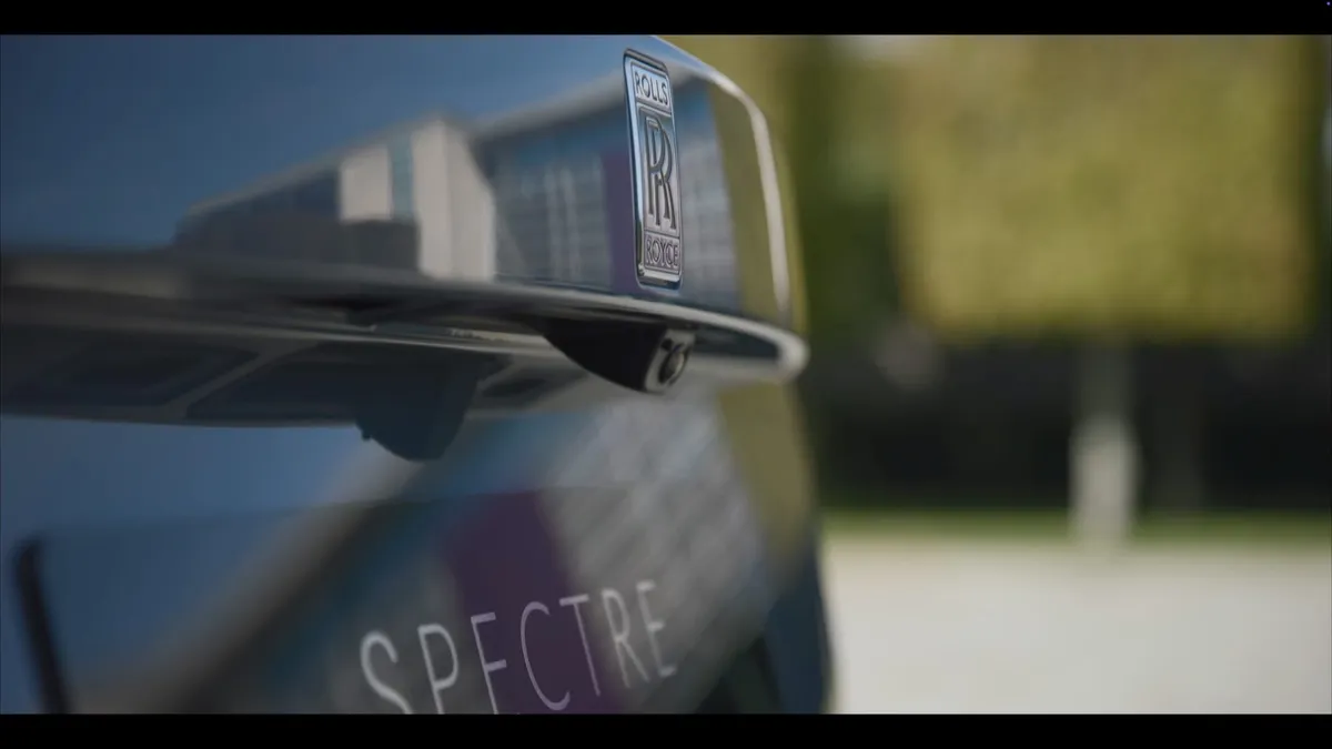 Close-up of a dark car's badge and side mirror mount, with "Spectre" lettering on the bonnet, buildings blurred in reflection.