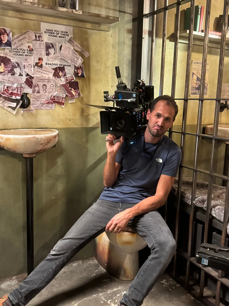 A camera operator in a blue T-shirt braces against a stool, filming with a shoulder-mounted camera in a set dressed as a jail cell with missing-persons posters on the wall.