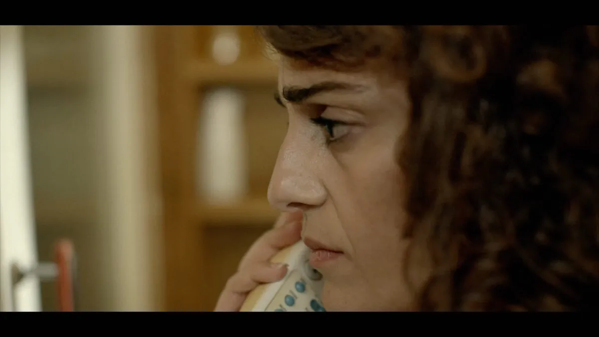 Close-up in profile of a curly-haired woman holding a corded telephone to her ear, a wood-panelled hallway blurred behind her.