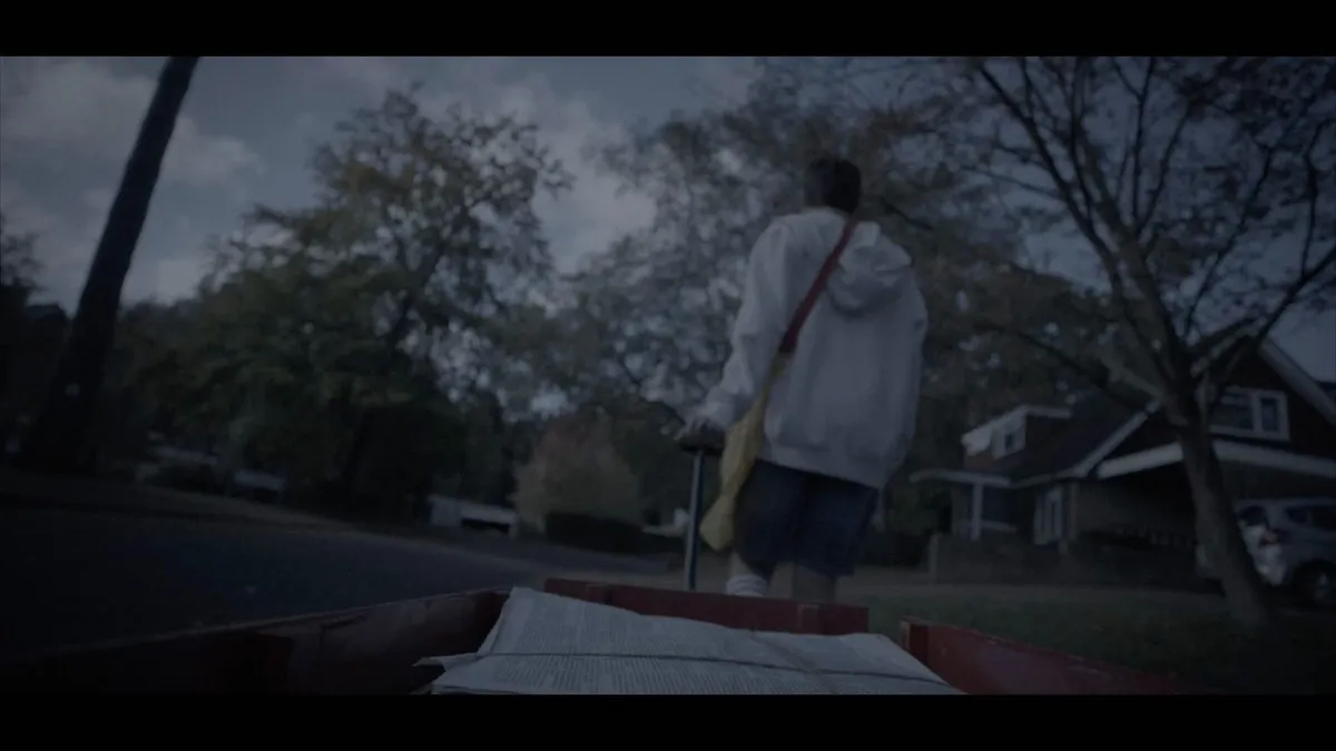 A boy in a white hoodie and denim shorts, seen from behind, pulls a wagon of newspapers along a suburban street at dusk.