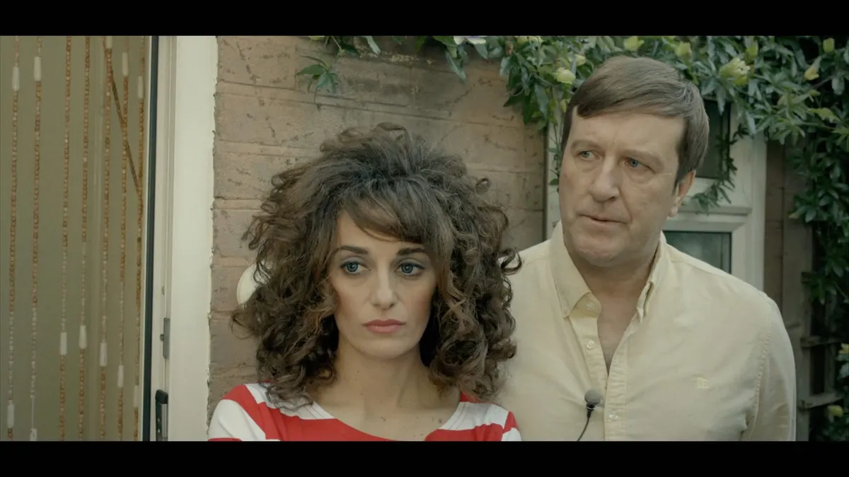 A curly-haired woman in a red-and-white striped top stands beside a man in a cream shirt, both outdoors near a brick wall and greenery.