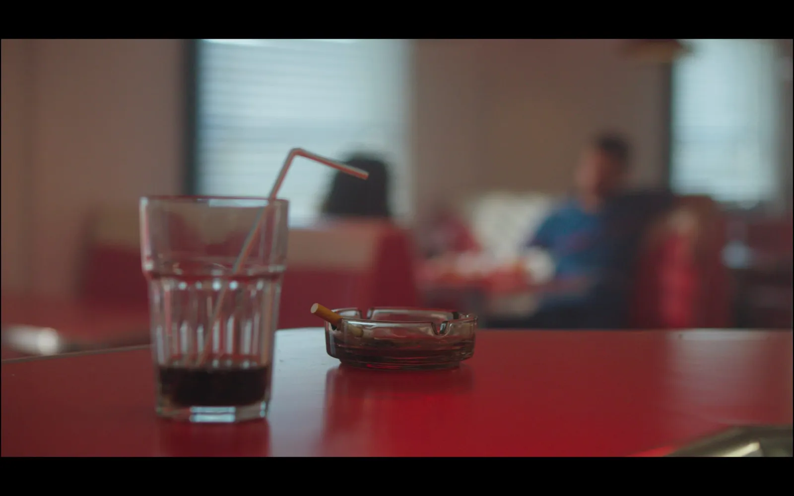 A glass of cola with a striped straw and a lit cigarette in an ashtray on a red diner table, figures blurred behind.