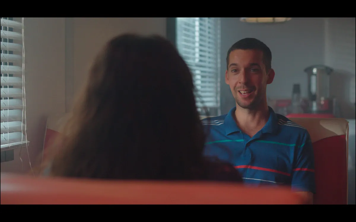 A man in a striped polo shirt smiles across a diner booth at a woman seen from behind, window blinds lit from outside.