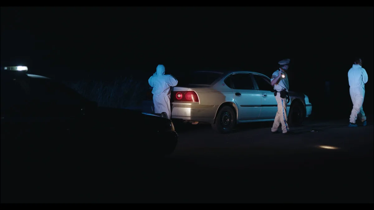 Night-time roadside scene with a saloon car lit by torchlight, an officer and two forensic crew in white overalls standing nearby.